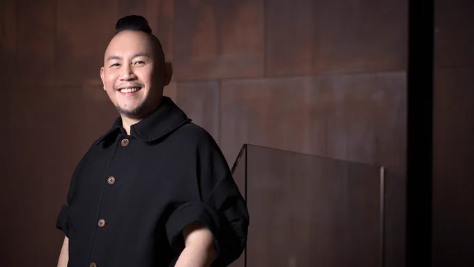 A half body shot of Low Kee Hong against a backdrop of a dark brown wall. Kee Hong is East Asian (Singaporean), and he is smiling widely at the camera, his body slightly facing the left. He is wearing a buttoned-up black shirt and is black hair is in a subtle mohawk.
