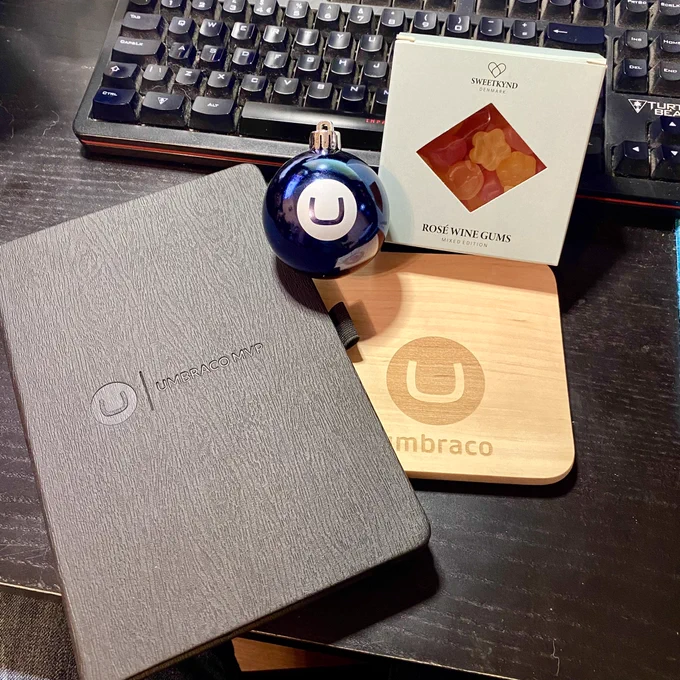 A black notebook, a small cheese board and bauble all with the Umbraco logo, alongside a box of fancy winegums, displayed on a black desk.