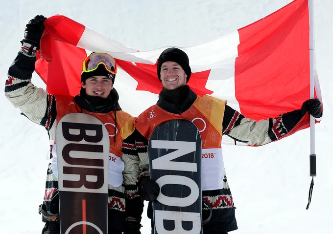 Two male athletes stand side by side holding up a Canadian flag behind them.