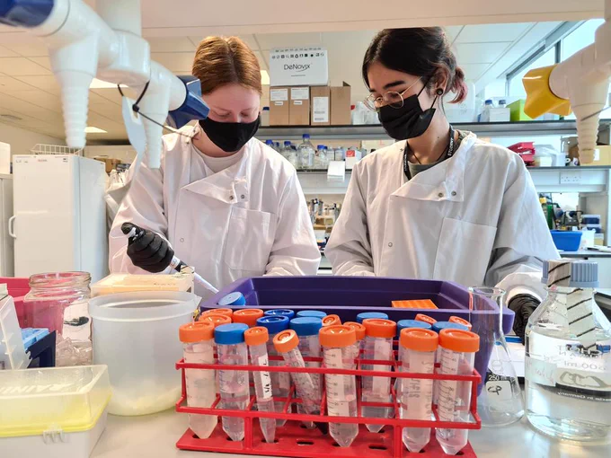 Photo graph of two students in a lab wearing lab coats and face masks , one is using a pipette.