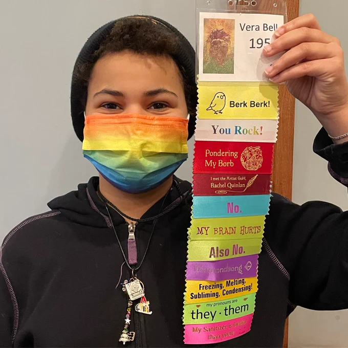Vera in a stocking cap holding up con ribbons including they/them pronouns.