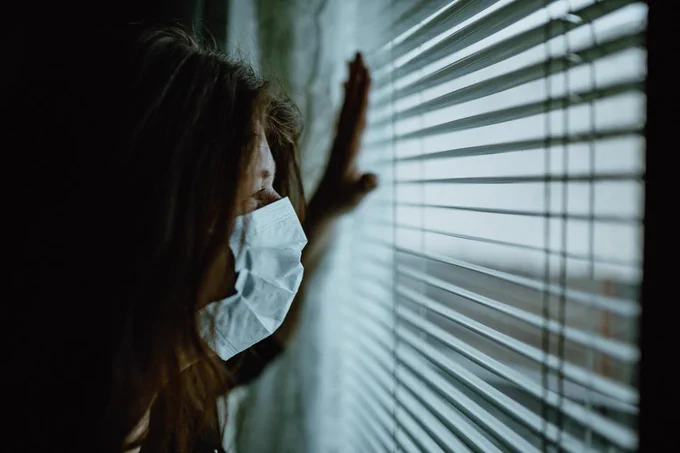 A masked person stares out of a window, through blinds.