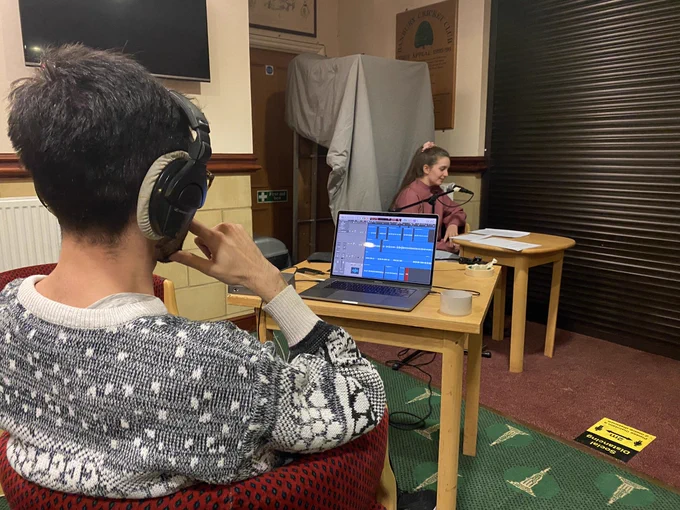 The photo looks over Amir’s shoulder towards Amelia sat at a table with a microphone.
