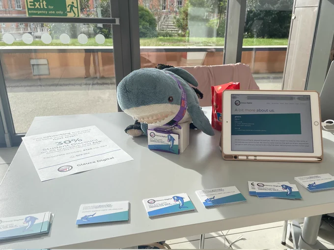 Photo of a table featuring a cute blue small plushy Blåhaj shark centre stage, surrounded by Glauca Digital flyers and business cards, and an iPad showing the Glauca website about page.