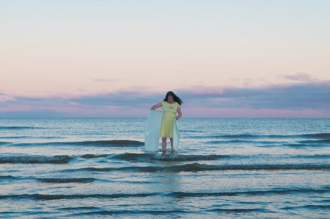 A woman looks as though she is standing on top of waves in a serene and beautiful environment, pink skies and blue calm ocean