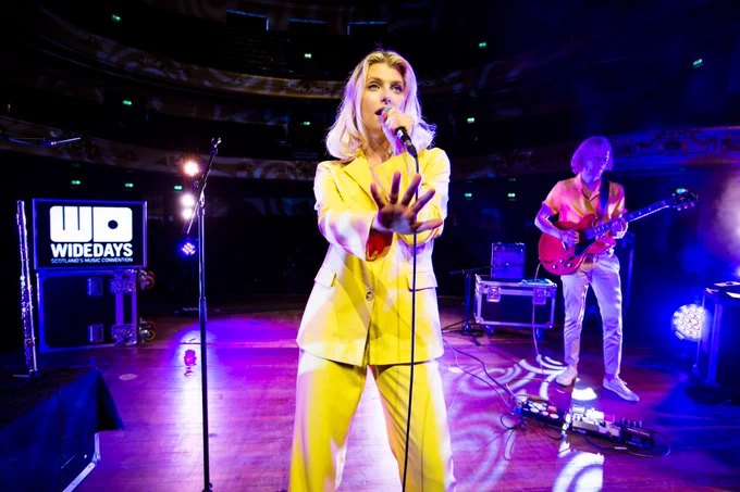 Solo artist Alex Amor performs at her showcase at Wide Days with a guitarist - she is wearing a yellow suit and reaches her hand out in front of her as she holds the microphone