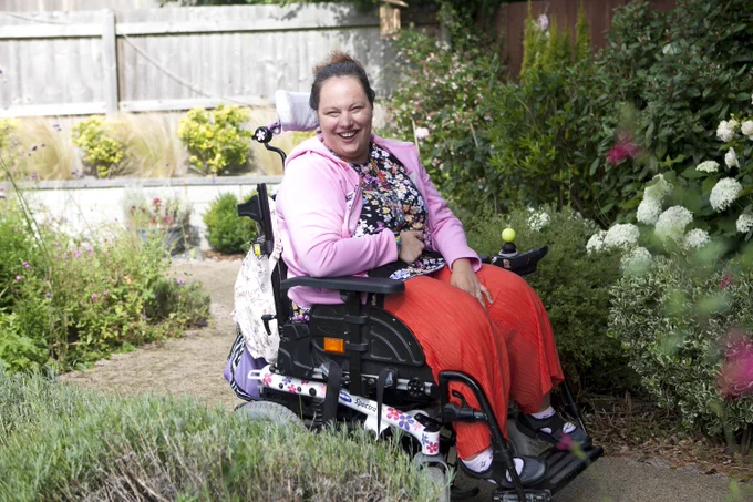 Image shows a woman in a powerchair in a garden. She is smiling and is wearing red trousers, a floral top and pink jumper.