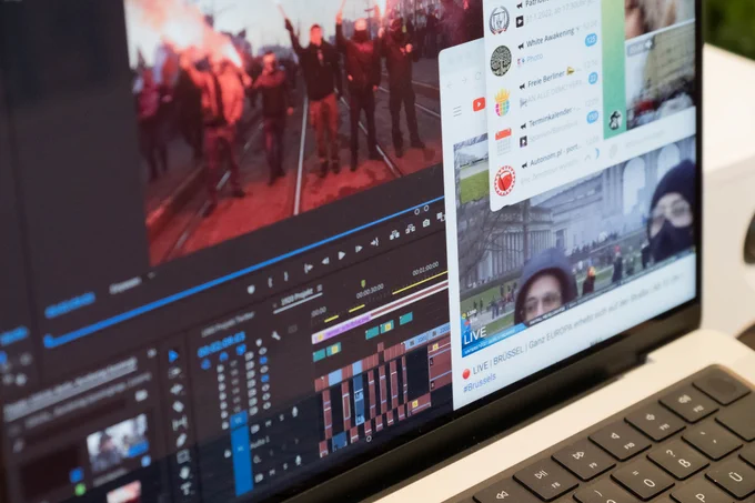 Foto von dem Bildschirm eines MacBooks, auf dem die Timeline eines Videoschnitt-Programms mit einem Standbild von Pyrotechnik, ein Fenster mit einem YouTube-Video von Protesten und ein Telegram-Chatfenster zu sehen sind.