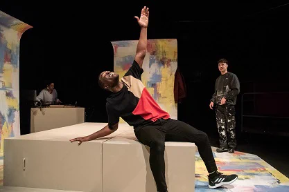 Justice Ritchie as Runaku and Neeta Sarl as the DJ in Human Nurture. Runaku is mid-speech and looks passionately and concerned. His arms are spread out and he reaches slightly upwards. The DJ is playing music in the background at a desk. 
