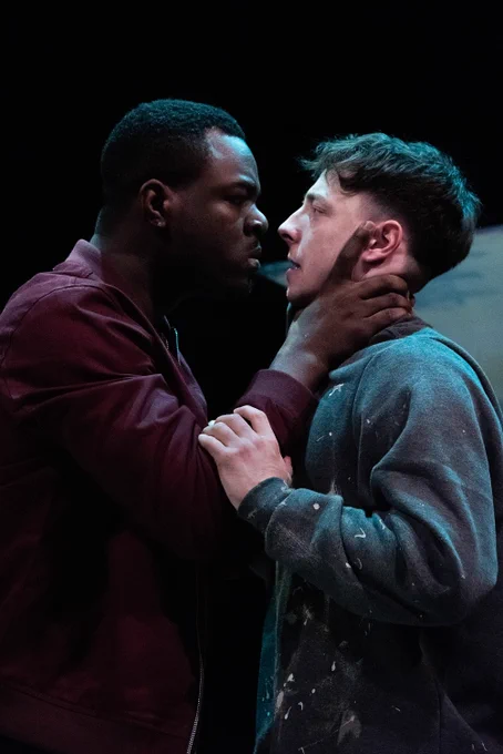 Justice Ritchie as Runaku and Lucas Button as Harry in Human Nurture. The men have been rowing, Runaku holds Harry by his neck and they both stand nose to nose. Runaku is angry, Harry looks scared and holds Runaku by the elbow