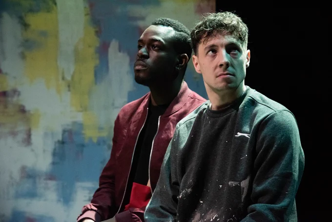 Justice Ritchie as Runaku and Lucas Button as Harry in Human Nurture. Both men sit next to one another, Runaku looks ahead blankly whilst Harry looks off to the side and upwards, as if something bad is about to happen. 