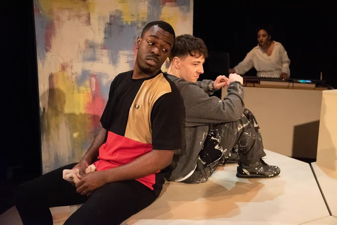 Justice Ritchie as Runaku, Lucas Button as Harry and Neeta Sarl as the DJ in Human Nurture. Runaku and Harry sit on a block, back to back, and lean over their shoulders to talk to one another. In the background, the DJ  is playing music at a desk.