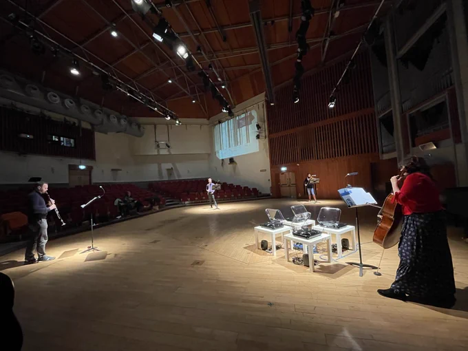 A performance with four musicians and four record players on stage. Clarinet, flute, violin and cello being played by four musicians standing up.