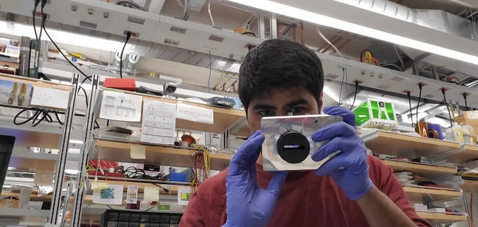 Photo of Kunal in the lab wearing nitrile gloves and holding up a rectangular device to their face