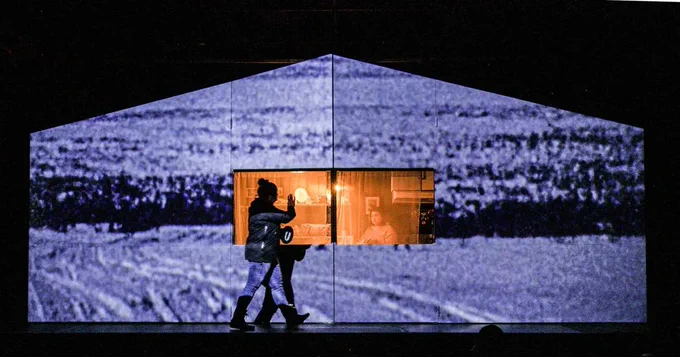 A scene from a play being performing on stage. A woman walks in front of a house and waves at someone through the window. 