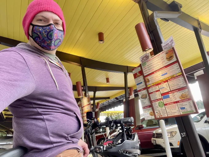 Wide angle selfie from waist level as I stand with my e-bike in a parking stall with fast food menu on display
