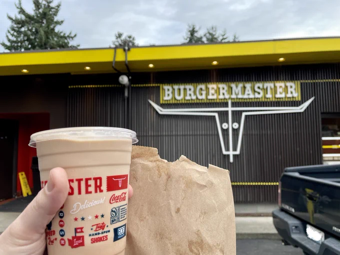 Hand holding a decorated plastic cup and brown paper bag with a sign reading “Burgermaster” over an abstract longhorn cow head