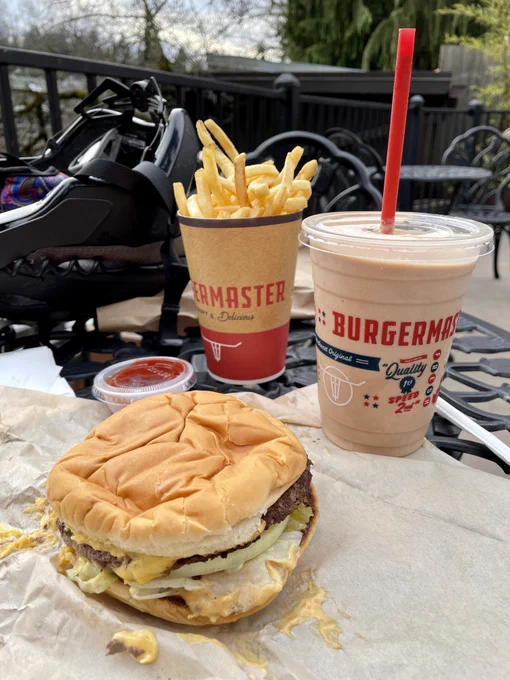 Cheeseburger with cheese, pickles, patty, and onion showing with a plastic cup & straw filled with chocolate milkshake and a cup filled with fries