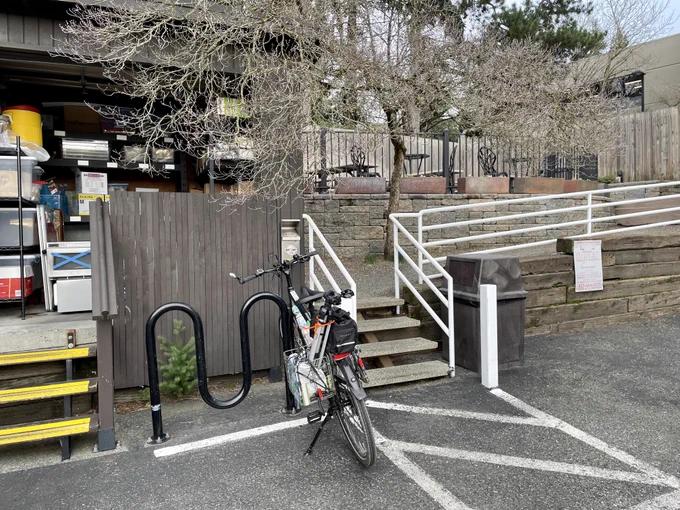 My e-bike leaning against a wave rack on the side of a parking lot with tables and chairs visible in an elevated patio