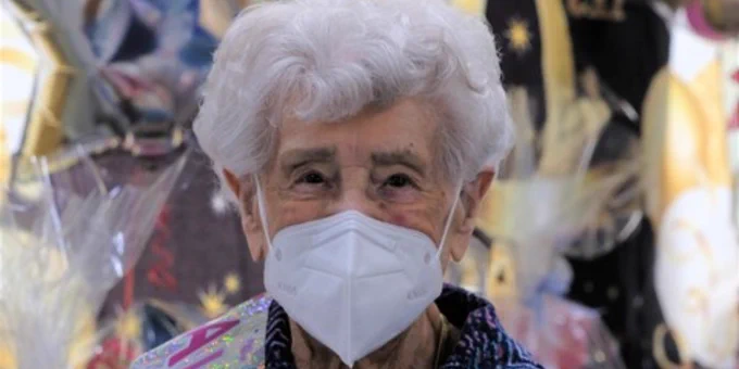Beryl is looking directly at the camera and holding a paper coffee cup. In the background is a 100th birthday balloon and Beryl is wearing a 'birthday girl' sash.