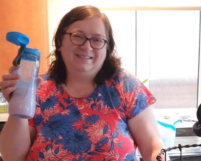 Karen is holding a shake up and smiling to camera. Karen is wearing a bright flowery top.