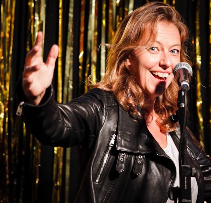 Rosie Wilby a white woman with shoulder length brown hair and a black leather jacker stands a microphone, arm outstretched, smiling