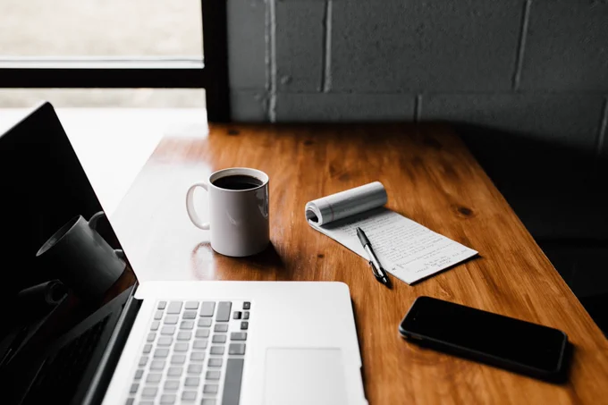 Laptop computer placed on a wooden table with a coffee mug, notepad, pen, and cell phone next to it.