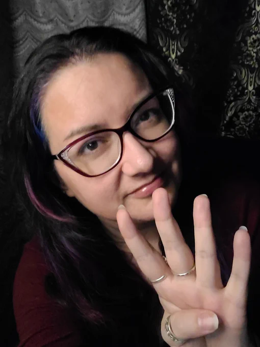 Middle-aged white woman, long brown/purple hair, catheter glasses, half smile. Hand extended partially in front of face, 4 fingers extended.