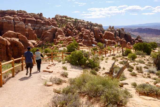 Two hikers walk along a fenced trail that overlooks the beige and orange striped spires of the Fiery Furnace on a bright, sunny day. Clouds are visible in the sky and trees, shrubs and stones line the trail in the foreground.