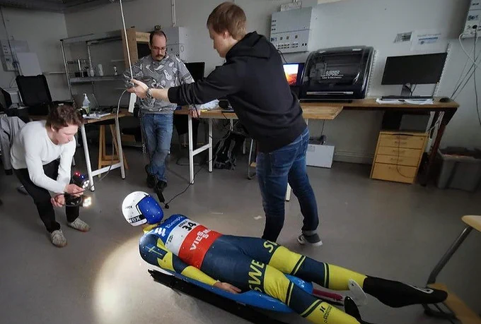 En person i tävlingsdräkt ligger ned på en rodel i kontorslokal med tre personer som omgärdar rodeln.