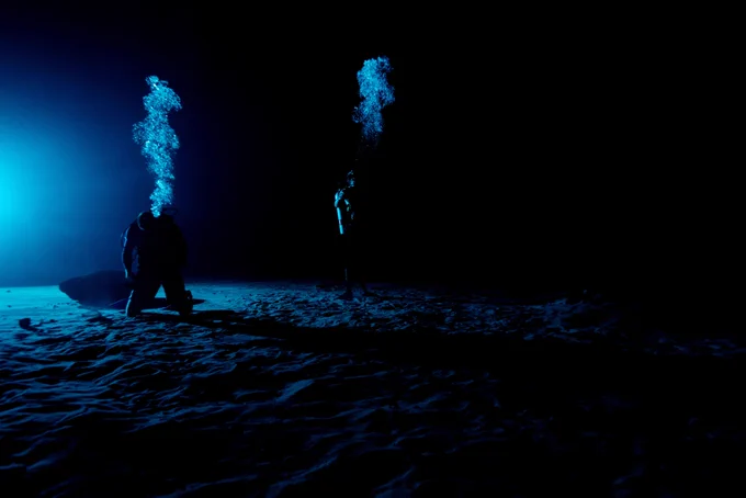 Two divers at the bottom of a swimming pool in the dark with two air bubbles emerging.