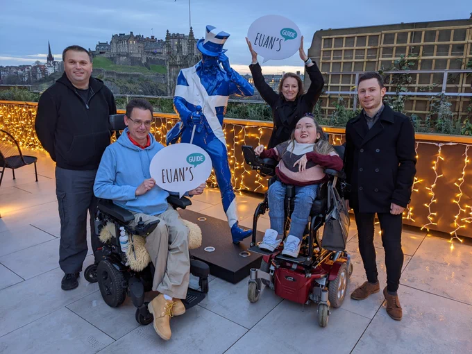 Five people next to a statue covered in the Scotland flag with two people holding the Euan's Guide logo.