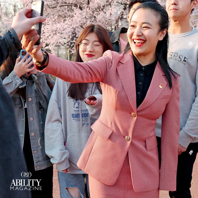 Chaofan in a pant suit smiling while holding up a phone, accompanied by 5 other people also smiling and holding their phones.