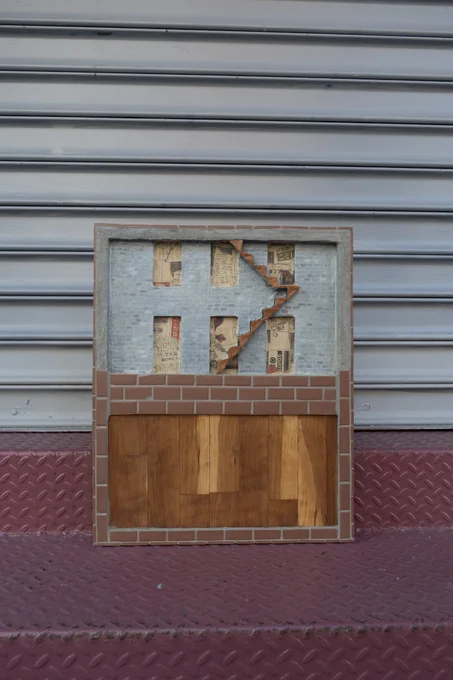 A rectangular panel sits on maroon steps against an industrial gray wall. The bottom half of the panel is wood surrounded by small bricks. The top top half is covered in small gray bricks. It has a small brown staircase and six window cutouts exposing newspaper.