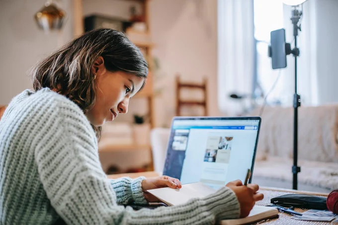 A young person with a blue sweater writing in a notebook. They are sitting in front of a laptop.