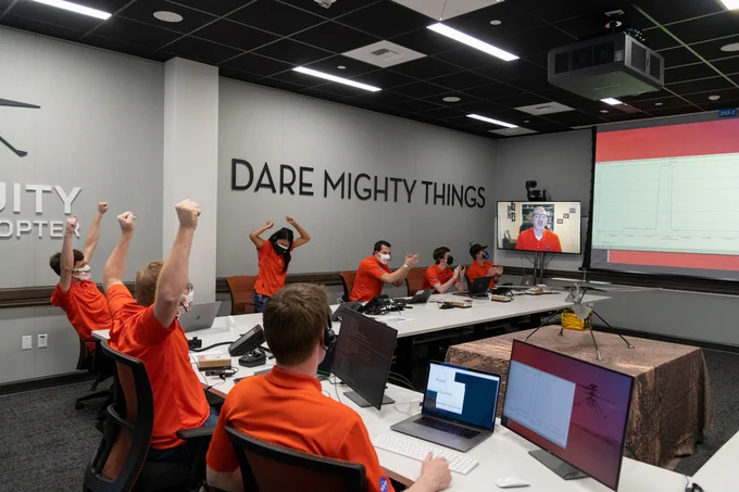 Group of people in orange polos sitting around a table cheer. On the wall are the words "Dare Mighty Things"