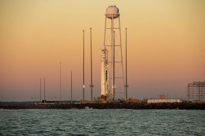 Antares rocket on pad, water in foreground and light pink sky in the background.