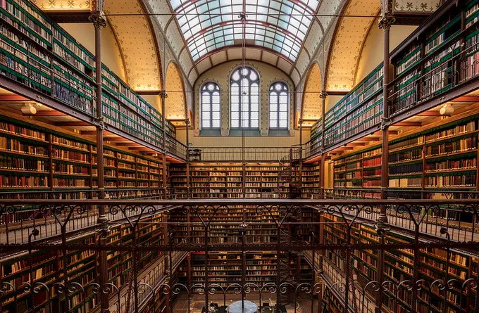 Photograph of the warmly-lit library inside the Rijks Museum in Amsterdam, Netherlands. An arched glass ceiling and three arched windows on the far wall, flanked by two walls of books at least four stories tall, girded by wrought iron railings and spiral staircases. 