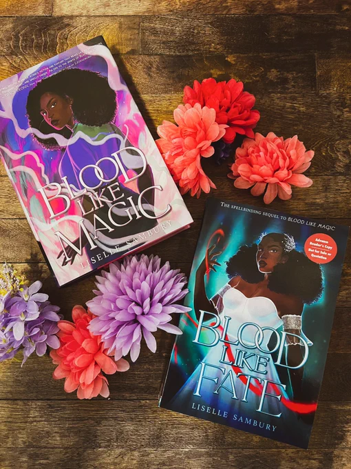 photo of Blood Like Magic and Blood Like Fate by Liselle Sambury on a hardwood floor with flowers around them