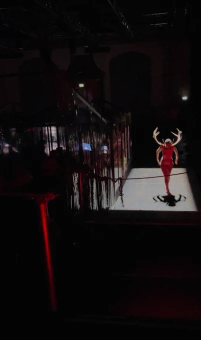 Inside a theatre, dark. On the left, a audience is sitting inside a greenhouse-like structure. On the right, a naked person walks naked carrying a large pair of deer antlers on their neck. The light is only on them, and creates a rectangular shape on the floor and reflections on the walls of the greenhouse