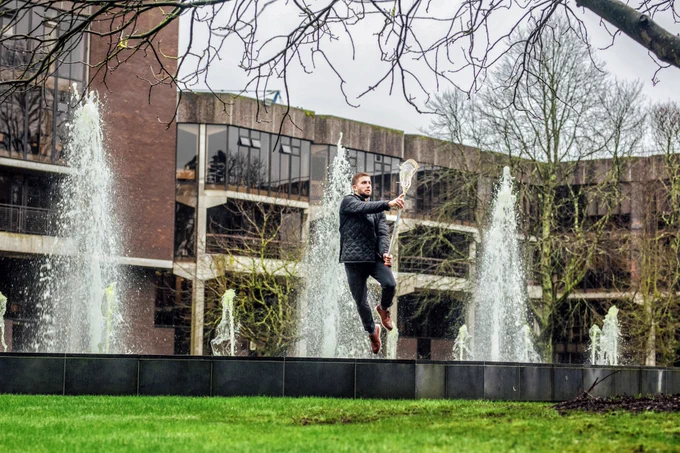 A man in a black padded jacket and dark coloured trousers, with brown shoes leaps into the air as he catches a ball in his lacrosse stick's net. He is stood on the ledge of a fountain, which is shooting up jets of water behind him. In the background are the main buildings of the University of Limerick