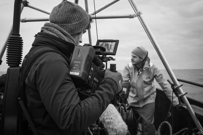 A man in the foreground looking into his camera monitor. Behind him a man stands at the edge of the boat.