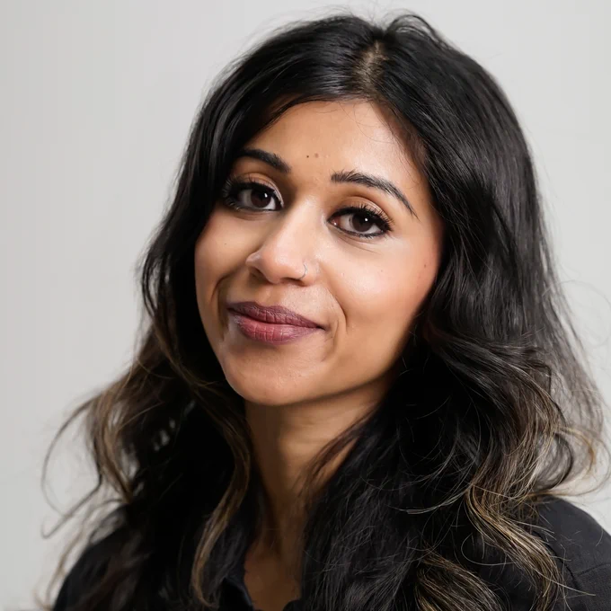 A headshot of Tamara Khandaker. Khandaker is on a grey background and slightly smiling to camera. Her hair goes past her shoulders and is dark brown.