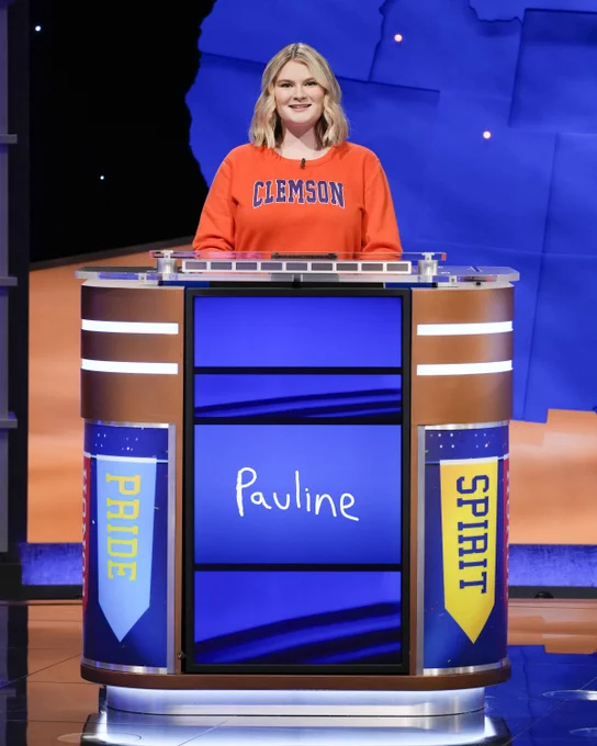 A woman wearing an orange shirt with Clemson across the front stands behind a Jeopardy podium. 