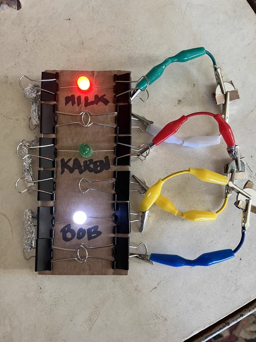 Board with led lights switches and batteries. Red and white lights are lit. Text says Mill Bob Karen.  An example of a Scrappy circuit