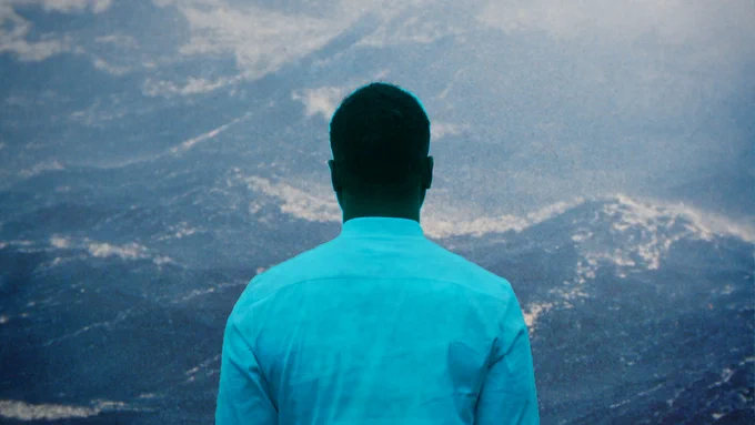 A still from "After Sherman." A person, tinted cyan and wearing a collared shirt, stares directly ahead at a hazy blue-gray nature-scape, back to the camera.