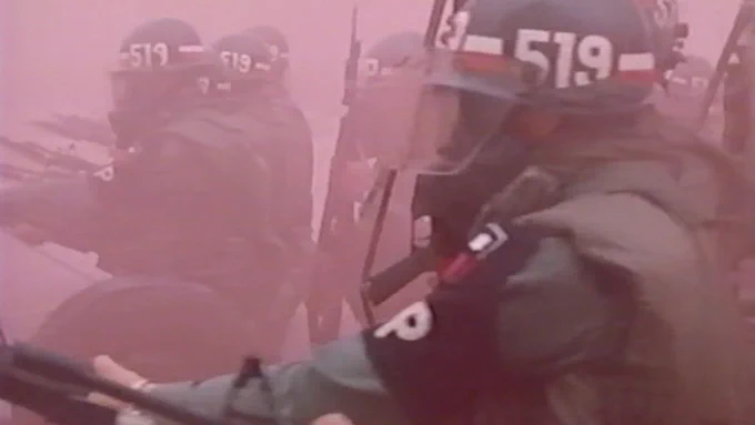 A still from "Riotsville, USA." A group of people wearing tactical gear in a pink haze.