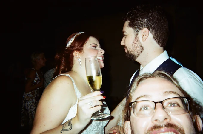 Selfie de um homem de óculos na frente de um casal de noivos prestes a se beijar. Ela usa um vestido branco e segura uma taça de champanhe.