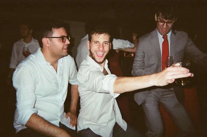 Três homens dançando. Dois estão de camisa branca, sorrindo, e o outro usa um terno cinza com gravata vermelha.