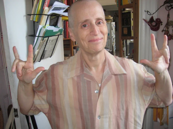 Leslie Feinberg smiles, making the ASL "I love you" sign with each hand, standing in office, with files and books.
Photo: Leslie Feinberg on 16th anniversary with Minnie Bruce Pratt, Jersey City, N.J., August 2008.
Credit: Minnie Bruce Pratt © 2008